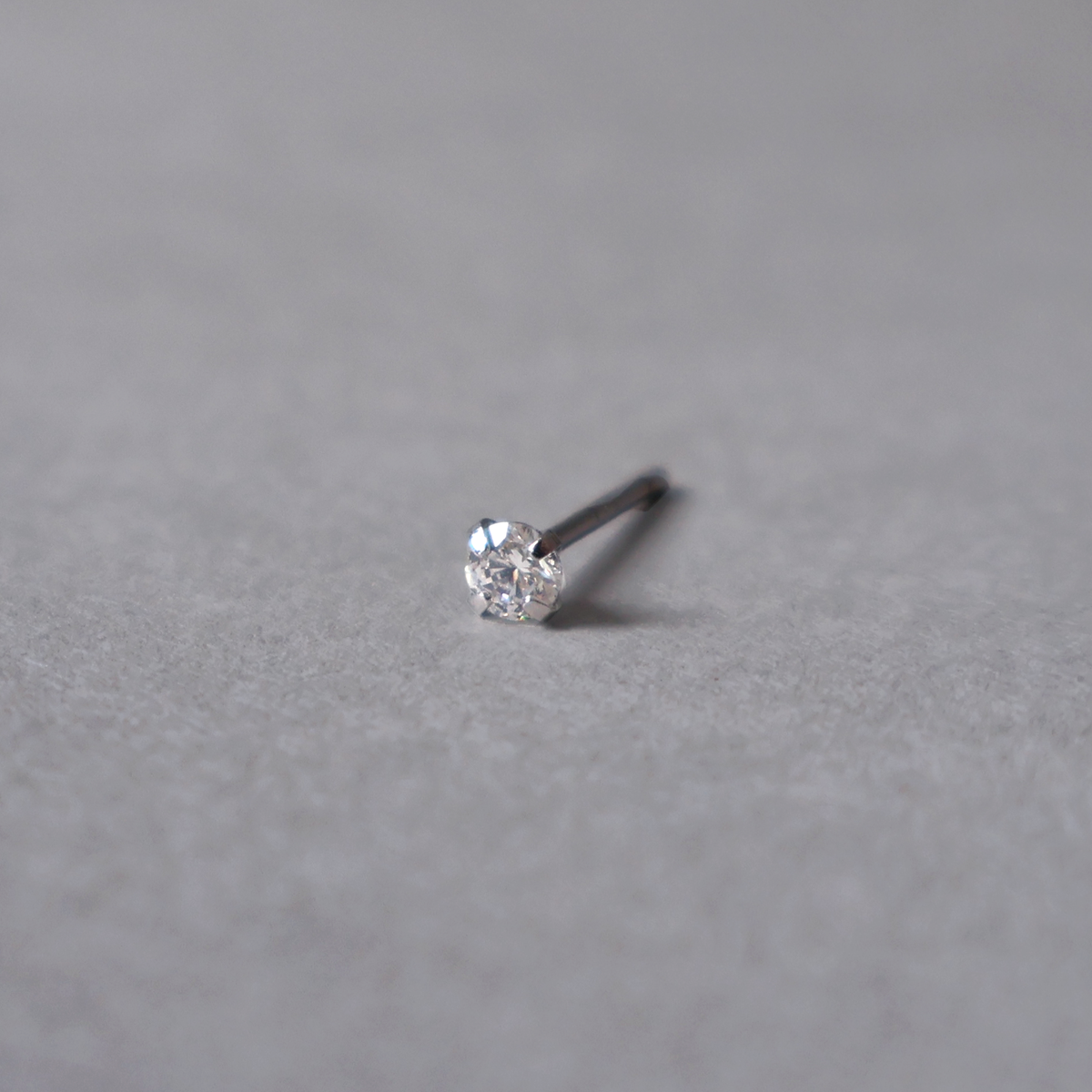 A single silver stud piercing featuring a round, brilliant-cut clear cubic zirconia gemstone held securely by four prongs, resting on a textured light-gray background.