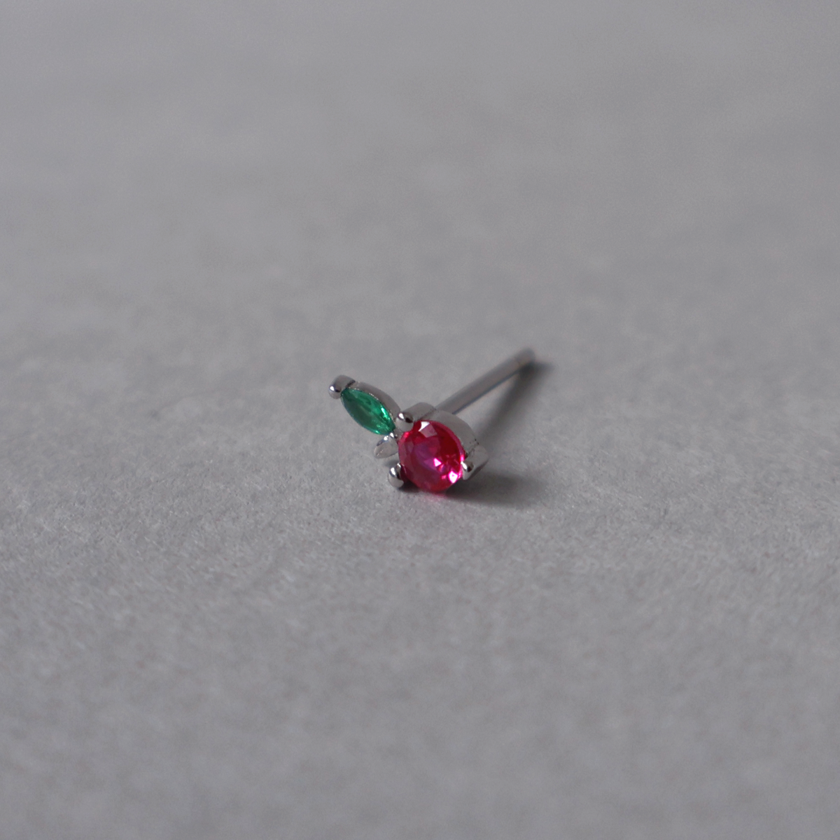Silver stud earring featuring a small apple design set with a round pink stone and a green leaf cubic zirconia. Detailed photo of the jewelry resting on a light neutral background.