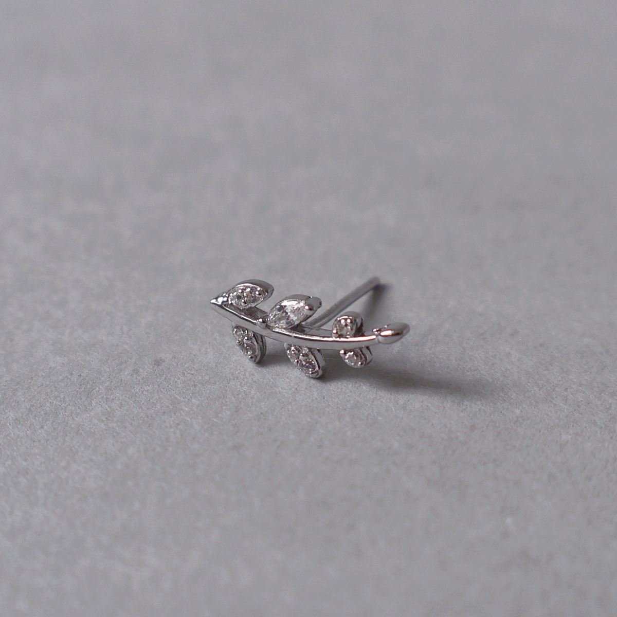 Silver stud earring shaped like a gently curved fern with leaves set with clear cubic zirconia. Detailed photo of the jewelry resting on a light neutral background.