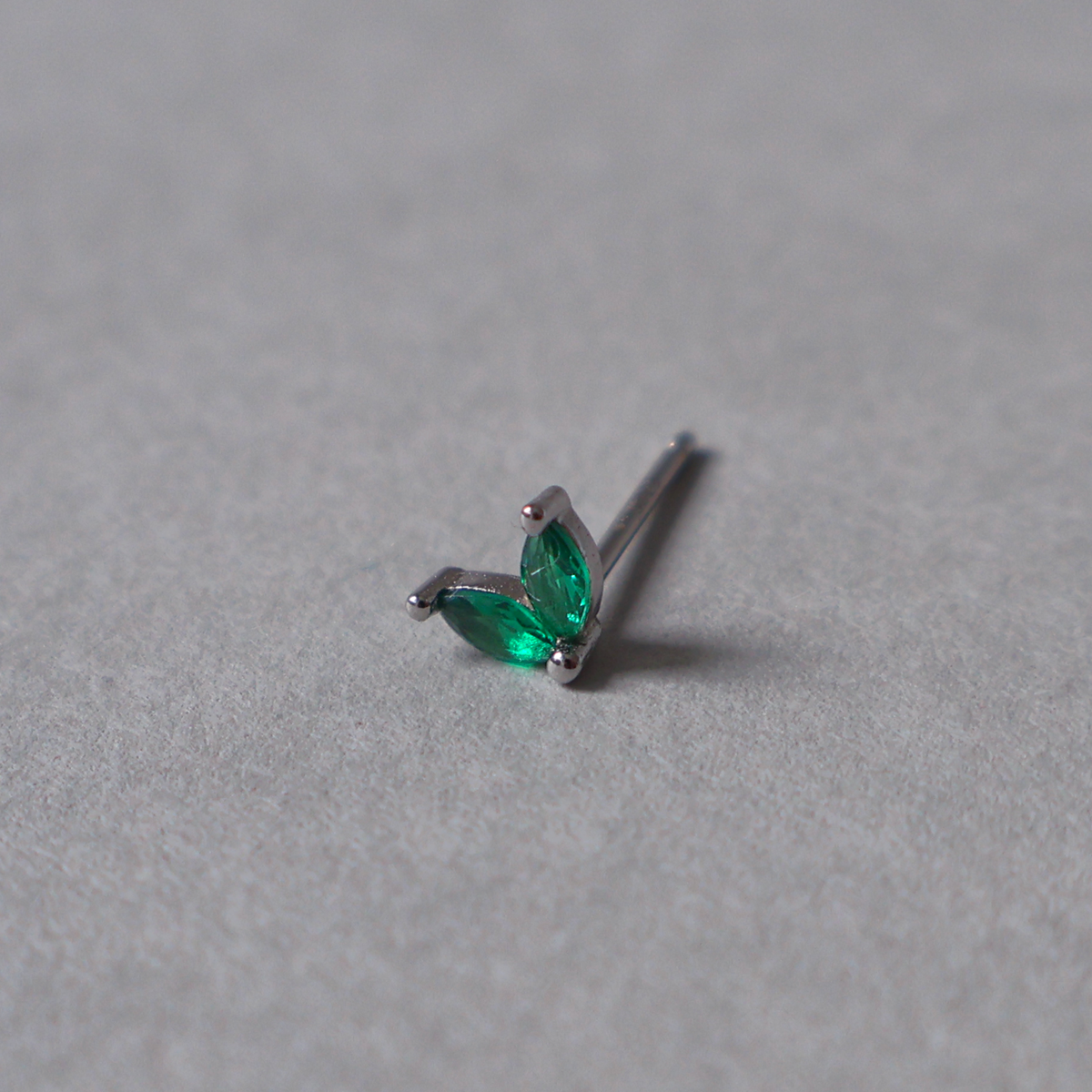 Silver stud earring featuring a delicate two-leaf design set with emerald green cubic zirconia. Detailed photo of the jewelry resting on a light neutral background.