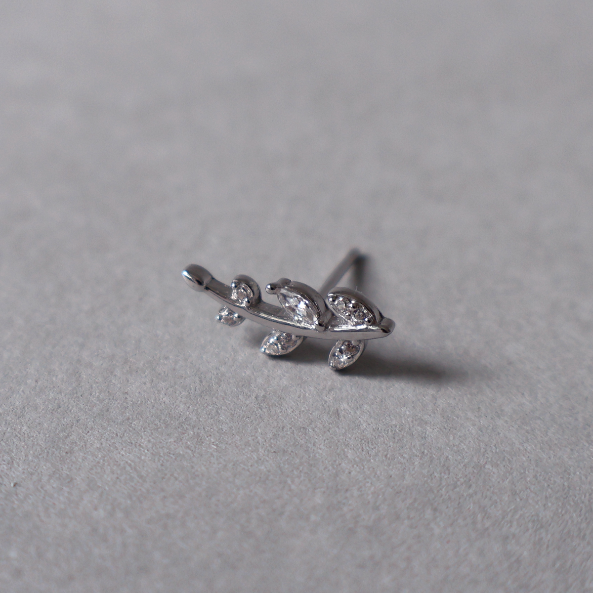 Silver stud earring shaped like a delicate curved branch with leaves set with clear cubic zirconia. Detailed photo of the jewelry resting on a light neutral background.