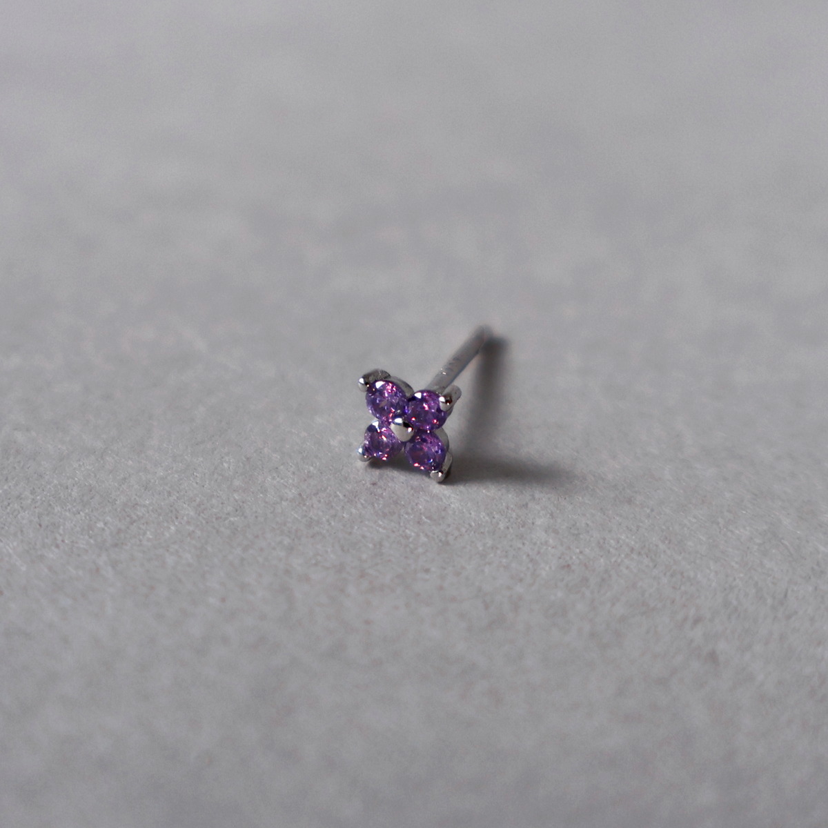 Silver stud earring featuring a small four-petal flower design set with purple cubic zirconia. Detailed photo of the jewelry resting on a light neutral background.